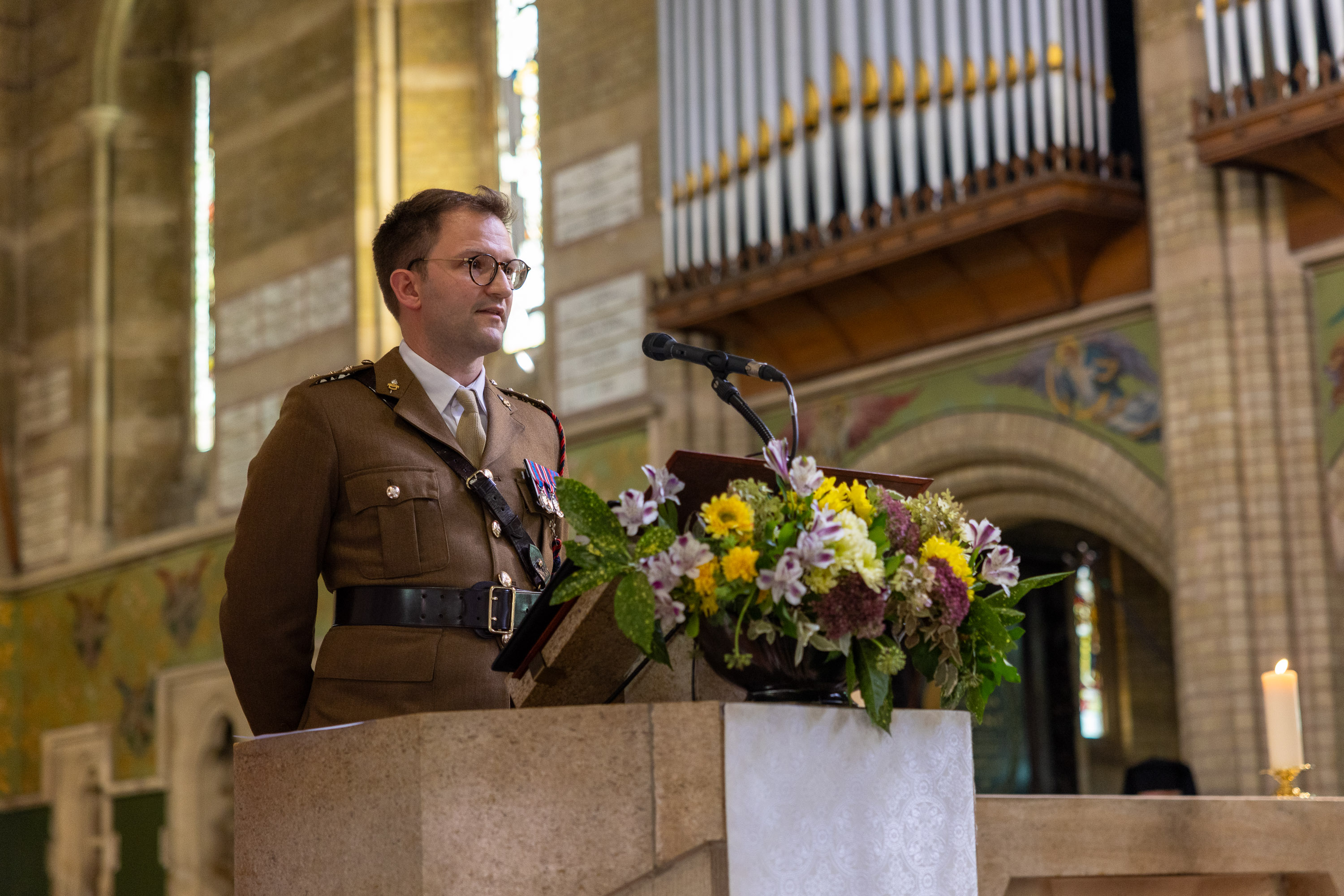 A military officer in uniform stands behind a stone lectern adorned with a colorful floral arrangement inside a church.