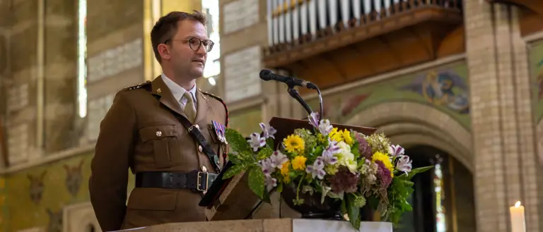 A military officer in uniform stands behind a stone lectern adorned with a colorful floral arrangement inside a church.