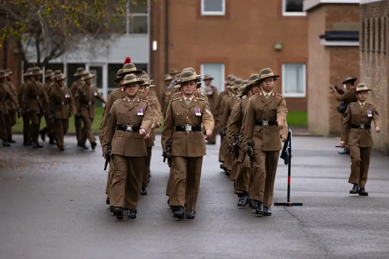 Soldiers in brown uniforms and brimmed hats march in formation on a grey street, with a brick building in the background.