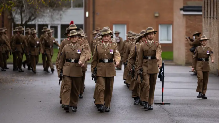 Soldiers in brown uniforms and brimmed hats march in formation on a grey street, with a brick building in the background.