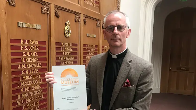A Vicar in formal attire and a clerical collar holds a certificate.