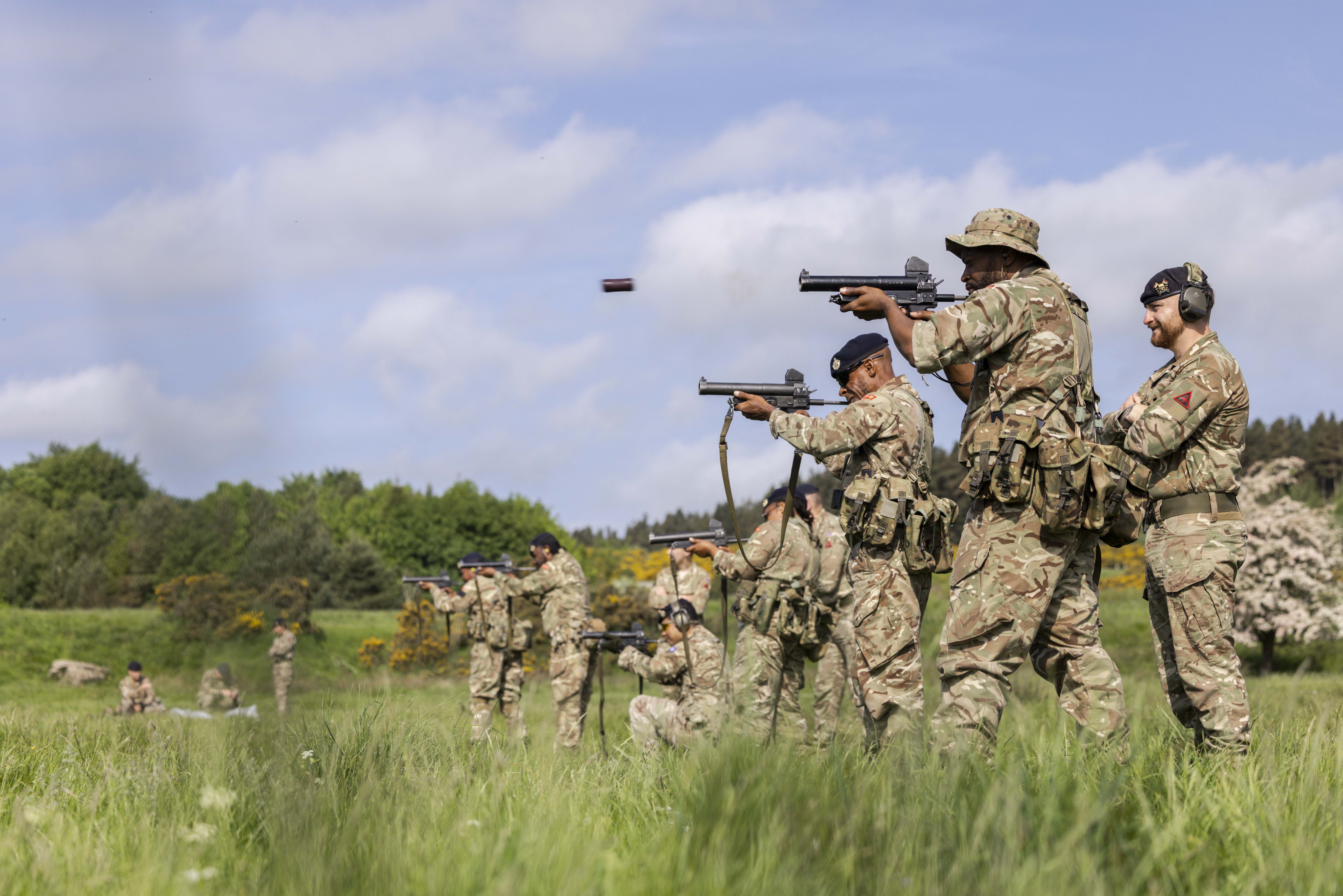 Soldiers in camouflage uniforms are training in a grassy field. They are firing towards targets.