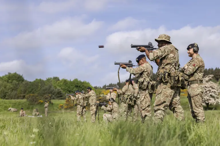 Soldiers in camouflage uniforms are training in a grassy field. They are firing towards targets.