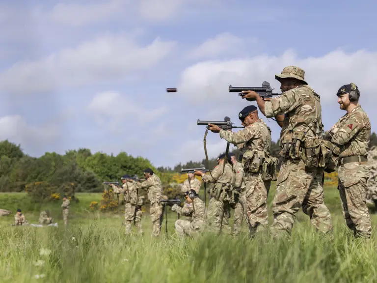 Soldiers in camouflage uniforms are training in a grassy field. They are firing towards targets.