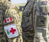 Two individuals in military uniforms stand side by side, facing each other. One uniform displays the Canadian flag and a Red Cross emblem, while the other features a British flag patch and an unidentified insignia. The background is an outdoor setting.