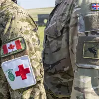 Two individuals in military uniforms stand side by side, facing each other. One uniform displays the Canadian flag and a Red Cross emblem, while the other features a British flag patch and an unidentified insignia. The background is an outdoor setting.
