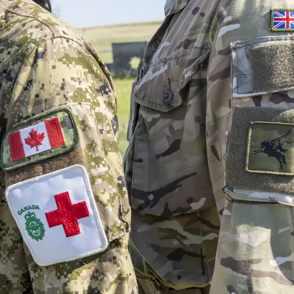Two individuals in military uniforms stand side by side, facing each other. One uniform displays the Canadian flag and a Red Cross emblem, while the other features a British flag patch and an unidentified insignia. The background is an outdoor setting.