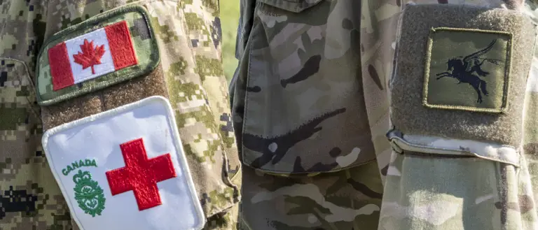 Two individuals in military uniforms stand side by side, facing each other. One uniform displays the Canadian flag and a Red Cross emblem, while the other features a British flag patch and an unidentified insignia. The background is an outdoor setting.