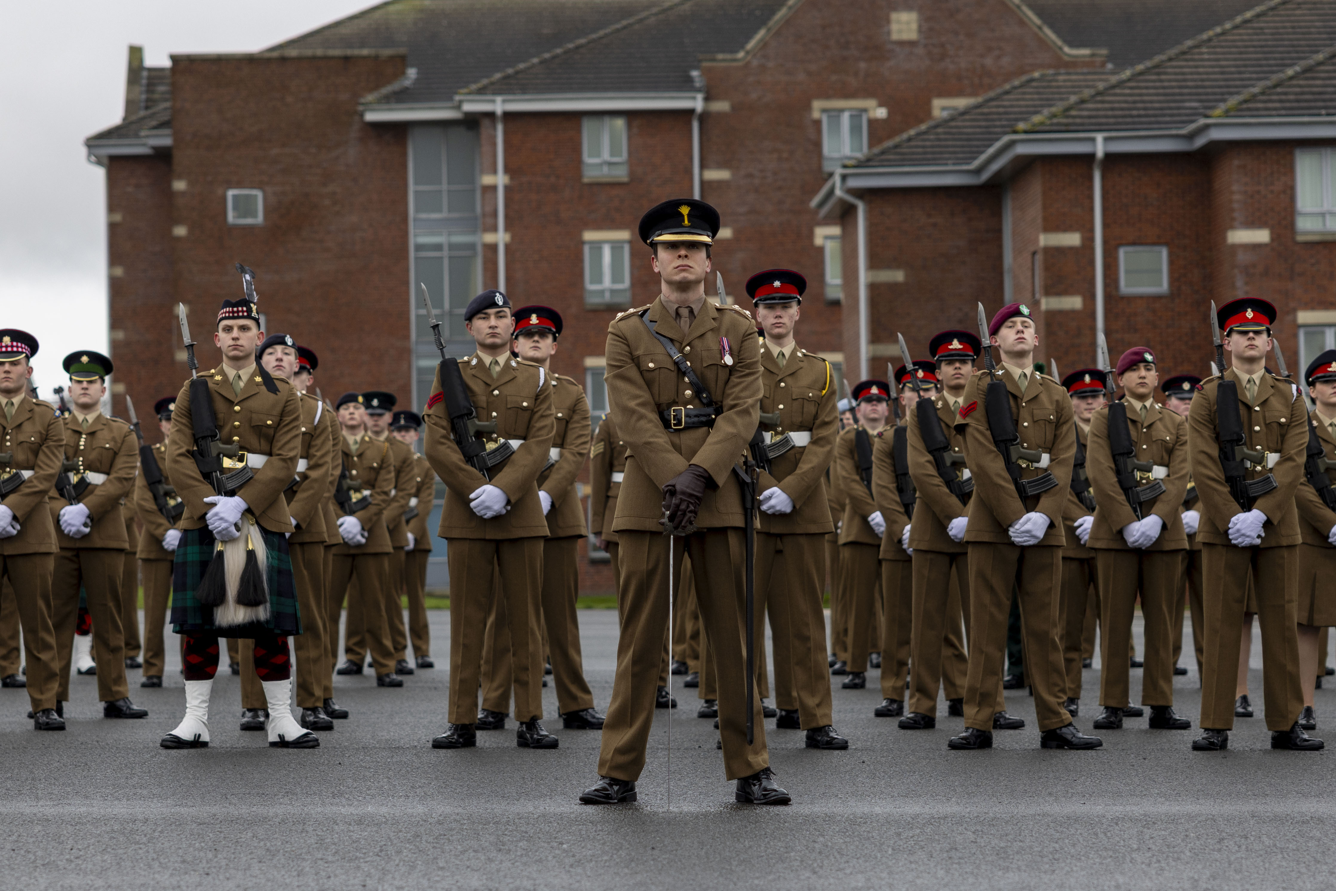 A group of uniformed soldiers stands in formation on a gray pavement against a backdrop of brick barracks. They are solemn and disciplined, exuding pride.