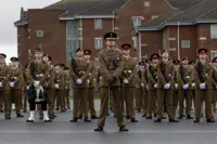 A group of uniformed soldiers stands in formation on a gray pavement against a backdrop of brick barracks. They are solemn and disciplined, exuding pride.