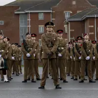 A group of uniformed soldiers stands in formation on a gray pavement against a backdrop of brick barracks. They are solemn and disciplined, exuding pride.