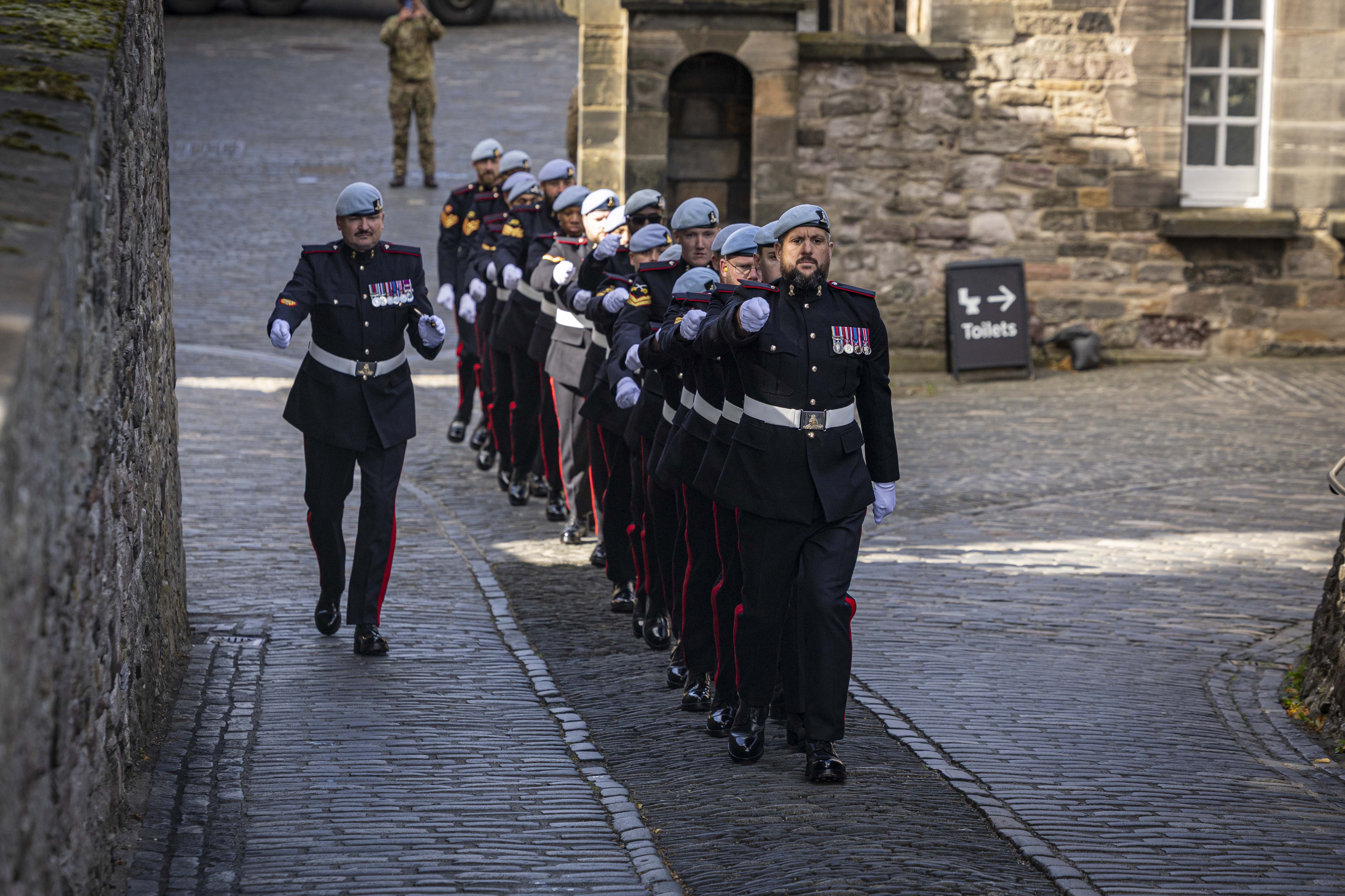 A group of uniformed guards is marching in formation along a cobblestone path. They're wearing dark uniforms with light blue berets and white gloves. The background features a historic stone building, and a sign pointing to toilets is visible. The setting is reminiscent of a ceremonial or official event.