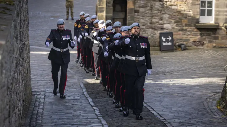A group of uniformed guards is marching in formation along a cobblestone path. They're wearing dark uniforms with light blue berets and white gloves. The background features a historic stone building, and a sign pointing to toilets is visible. The setting is reminiscent of a ceremonial or official event.