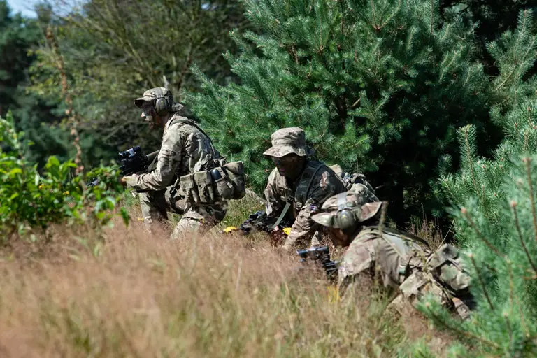 Three individuals in camouflage uniforms crouch among tall grasses and trees, holding rifles and wearing helmets with goggles. The setting appears to be a forested area.