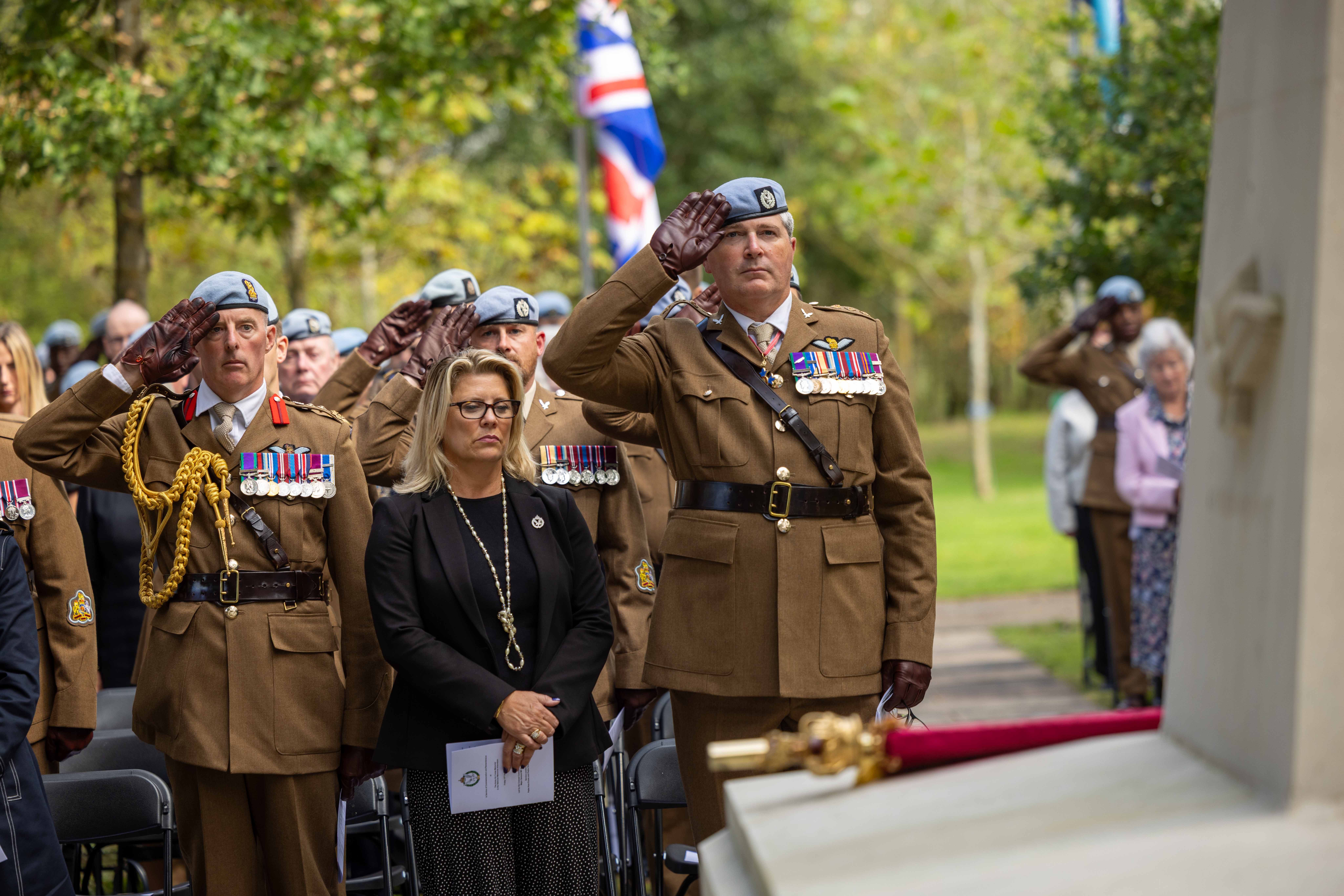 A group of military personnel in uniform and a civilian stand at attention during a ceremonial event. Some are saluting near a memorial with flags, including the United Kingdom’s flag, visible in the background. The setting is outdoors with trees nearby.