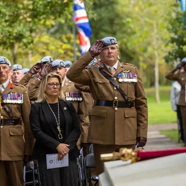 A group of military personnel in uniform and a civilian stand at attention during a ceremonial event. Some are saluting near a memorial with flags, including the United Kingdom’s flag, visible in the background. The setting is outdoors with trees nearby.