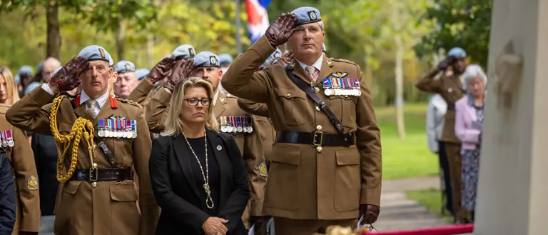 A group of military personnel in uniform and a civilian stand at attention during a ceremonial event. Some are saluting near a memorial with flags, including the United Kingdom’s flag, visible in the background. The setting is outdoors with trees nearby.
