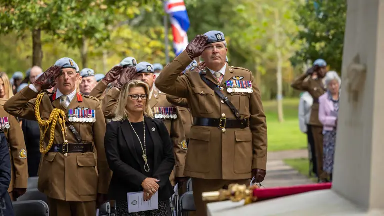 A group of military personnel in uniform and a civilian stand at attention during a ceremonial event. Some are saluting near a memorial with flags, including the United Kingdom’s flag, visible in the background. The setting is outdoors with trees nearby.