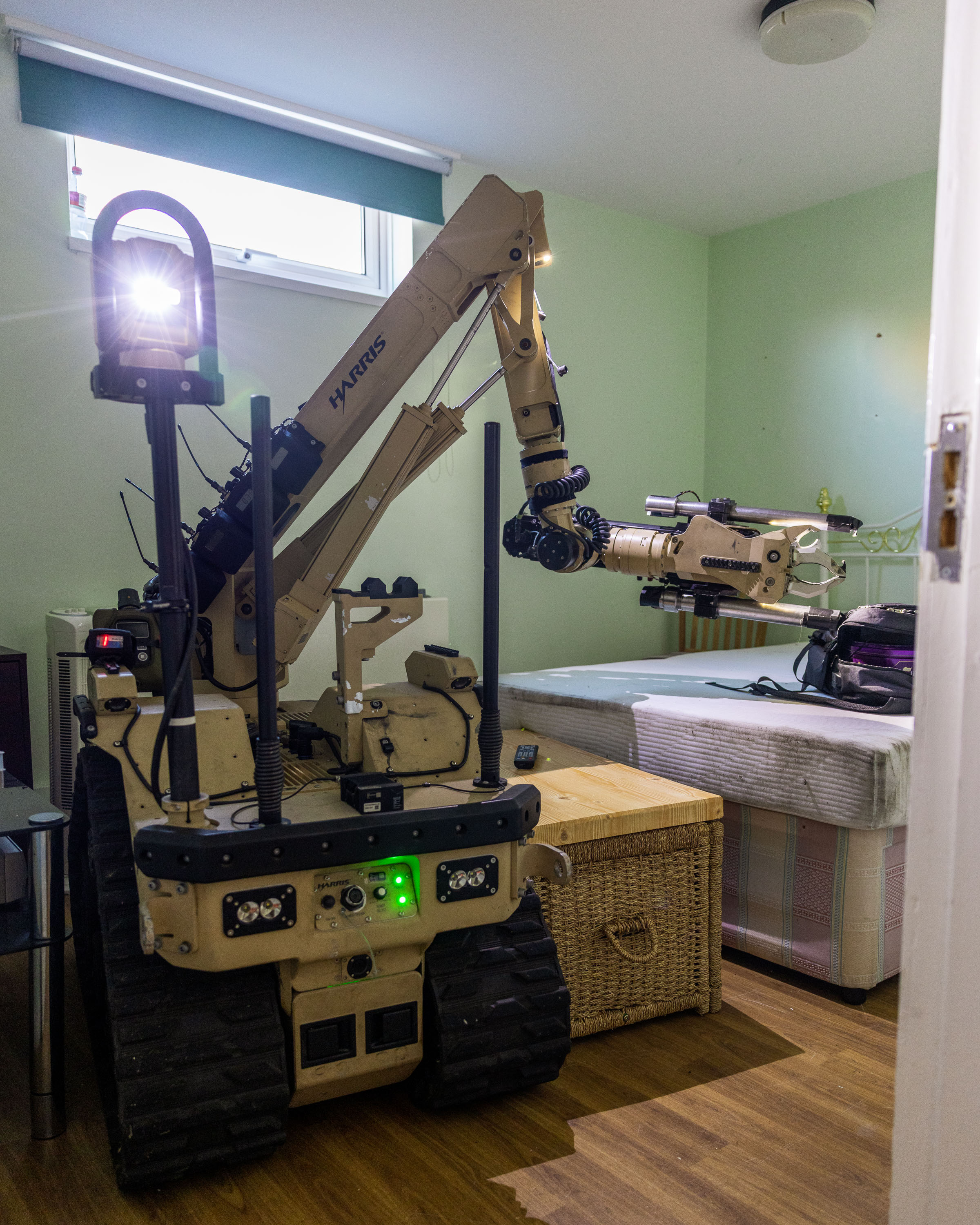 A sand coloured bomb disposal robot sits in a room. It is looking at a bag on a bed.