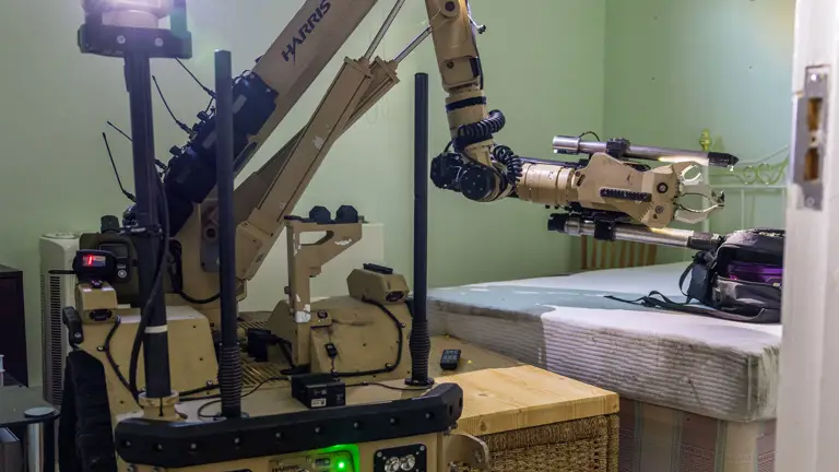 A sand coloured bomb disposal robot sits in a room. It is looking at a bag on a bed.
