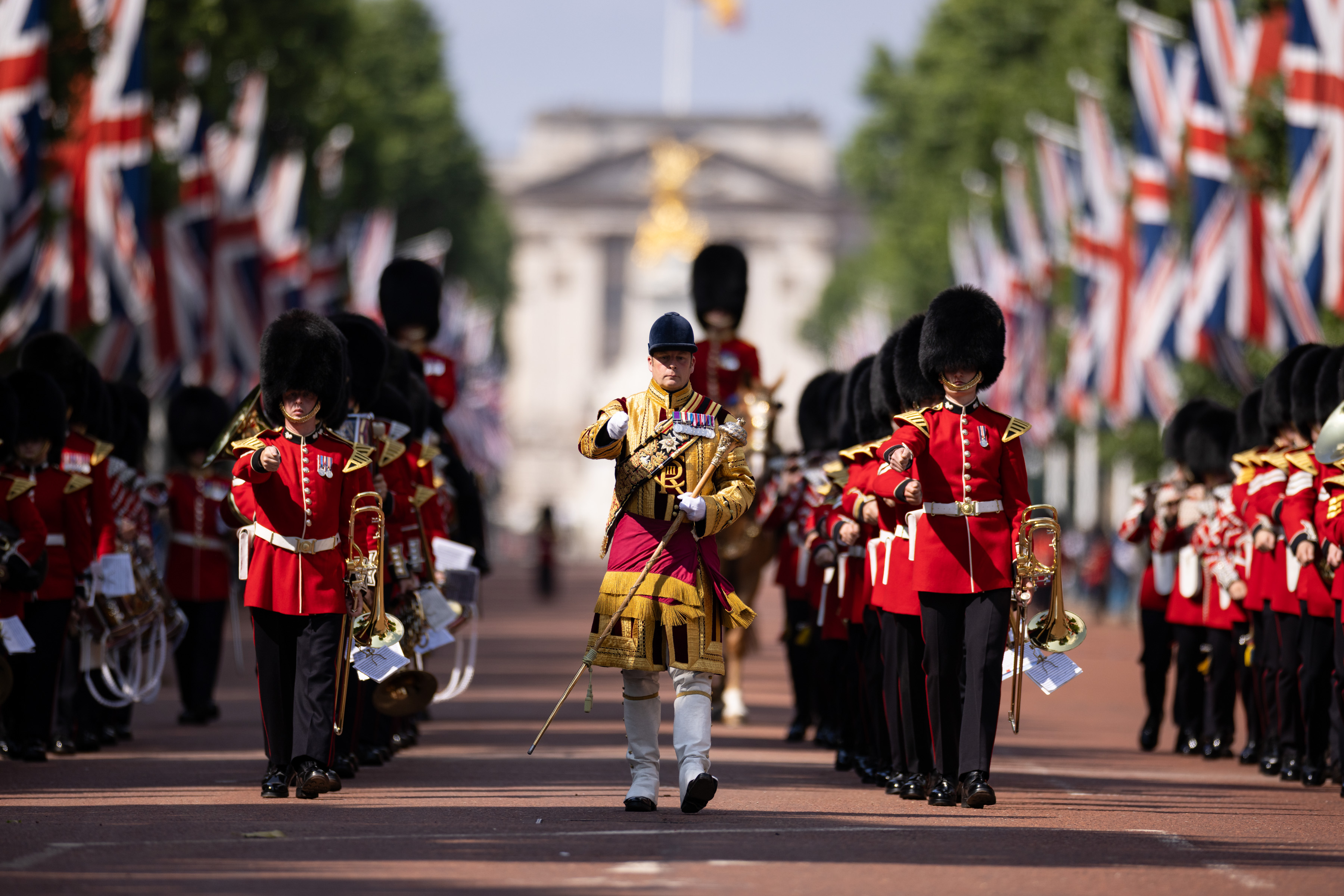 Four lines of soldiers in red ceremonial attire march behind a soldier in yellow ceremonial attire.