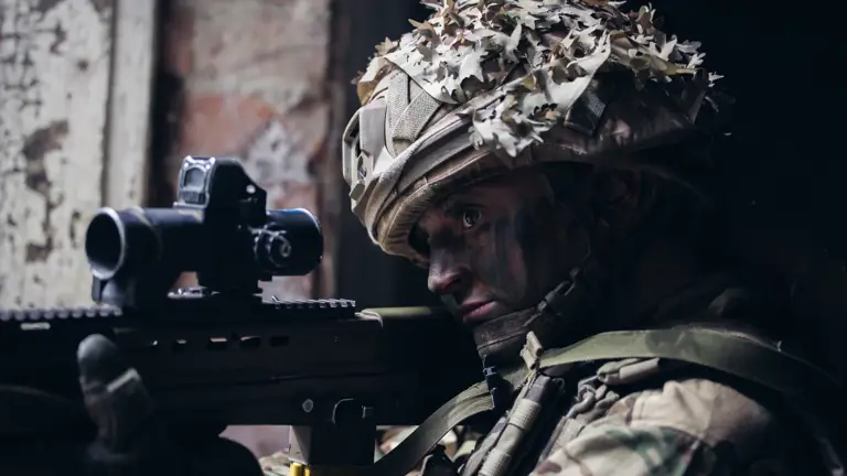 A soldier takes aim with her weapon, pointing it outside from a window.