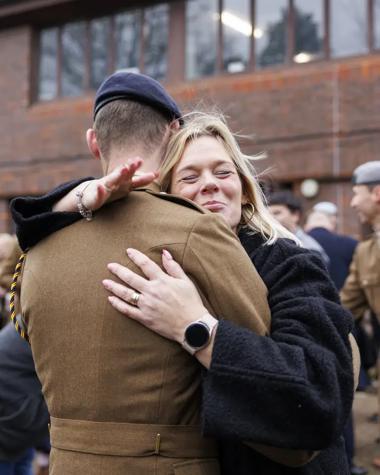 A person in a military uniform receives a heartfelt hug from another person wearing a dark coat in an outdoor setting.