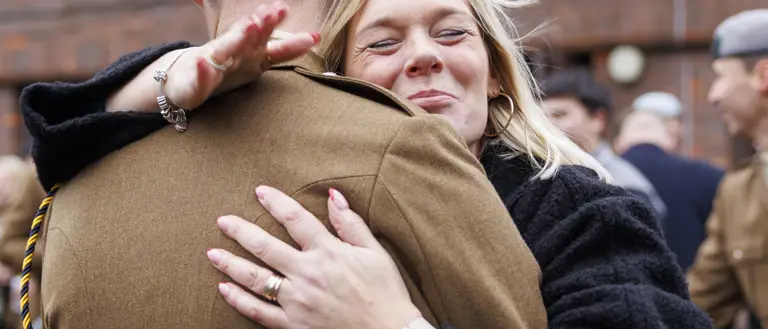 A person in a military uniform receives a heartfelt hug from another person wearing a dark coat in an outdoor setting.