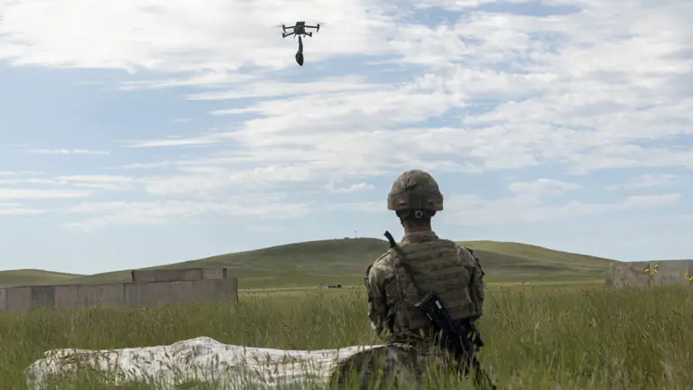 A person in military uniform is seated on a grassy field, observing a flying drone in the sky. There is a small hill and some concrete structures in the background under a cloudy blue sky.