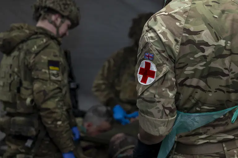 Military personnel are tending to a patient in a medical tent. The focus is on uniforms with camouflage patterns.