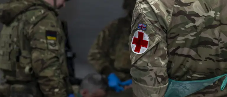 Military personnel are tending to a patient in a medical tent. The focus is on uniforms with camouflage patterns.