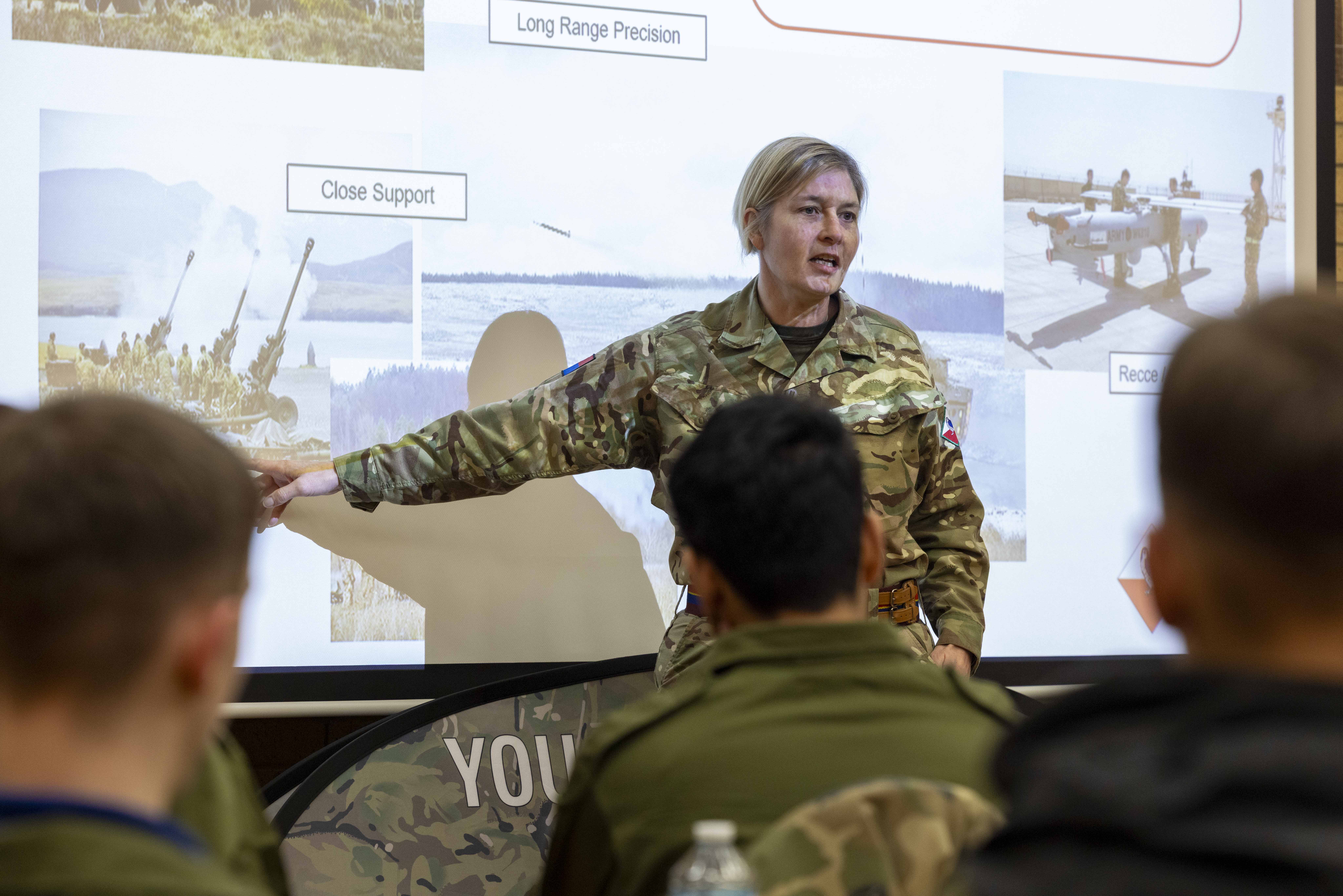 Military instructor in camouflage uniform pointing at a presentation slide.
