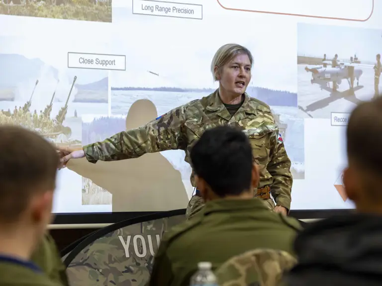 Military instructor in camouflage uniform pointing at a presentation slide.
