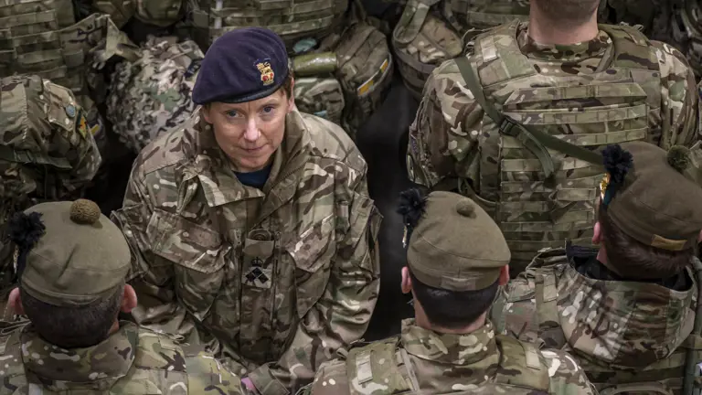 An officer in uniform is pictured facing the camera, she is positioned in the middle of a crowd of other soldiers, who are facing the other way.