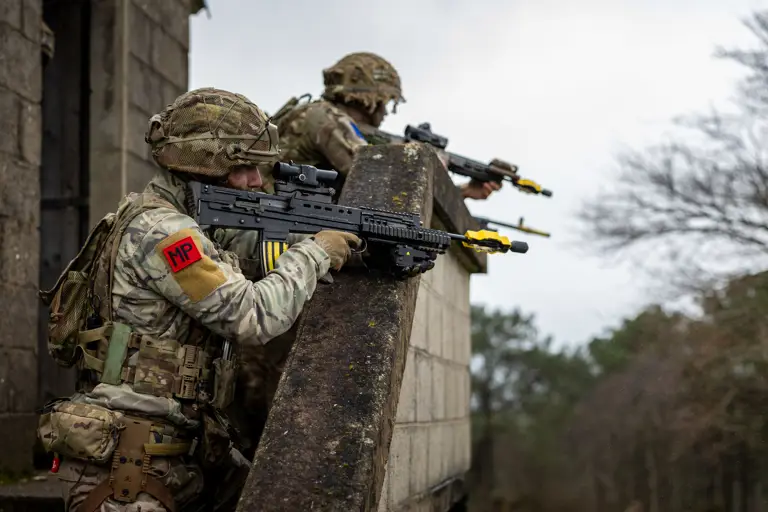 Two soldiers in camouflage gear and helmets are leaning over a stone wall. They are holding rifles with yellow-tipped barrels. One soldier has an