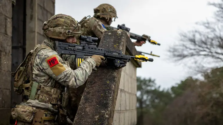 Two soldiers in camouflage gear and helmets are leaning over a stone wall. They are holding rifles with yellow-tipped barrels. One soldier has an