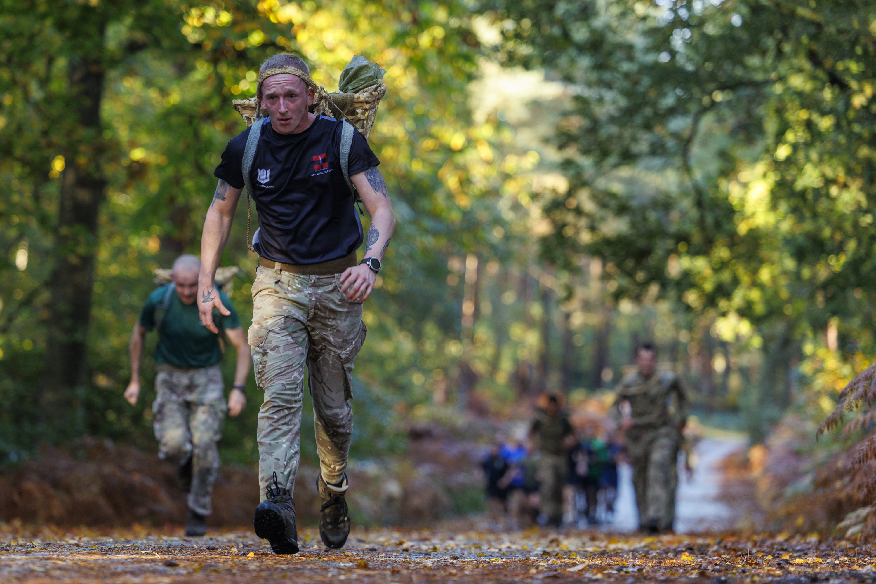 A soldier in camouflage trousers and a black shirt carries a heavy backpack while running on a forest trail.