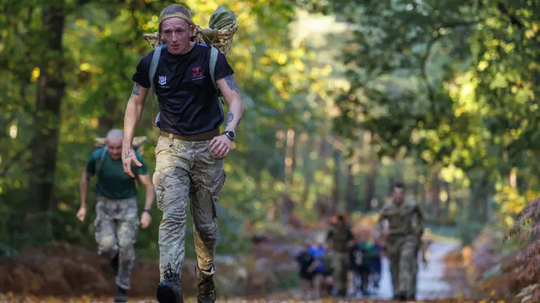 A soldier in camouflage trousers and a black shirt carries a heavy backpack while running on a forest trail.