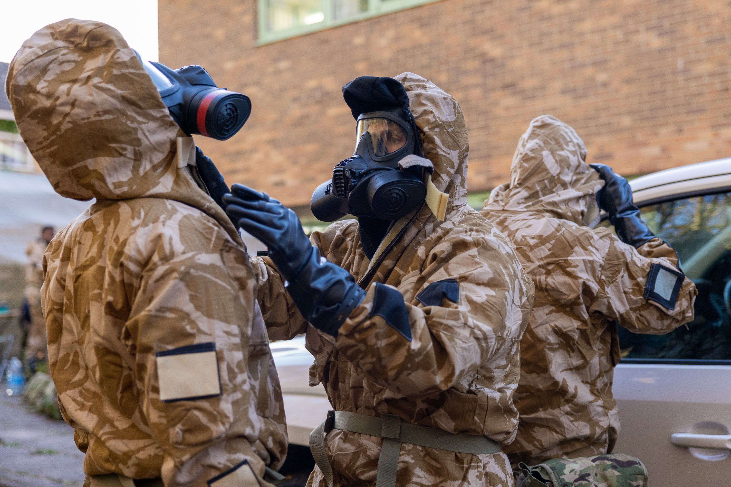 Three individuals in camouflage hazmat suits and gas masks prepare for a hazardous environment near a vehicle.