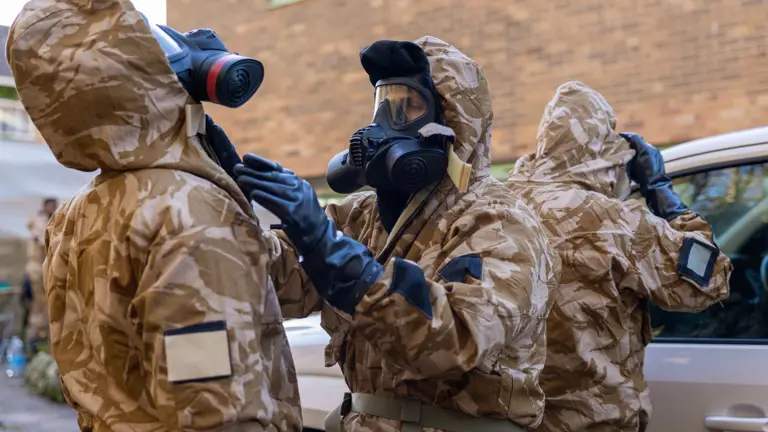 Three individuals in camouflage hazmat suits and gas masks prepare for a hazardous environment near a vehicle.