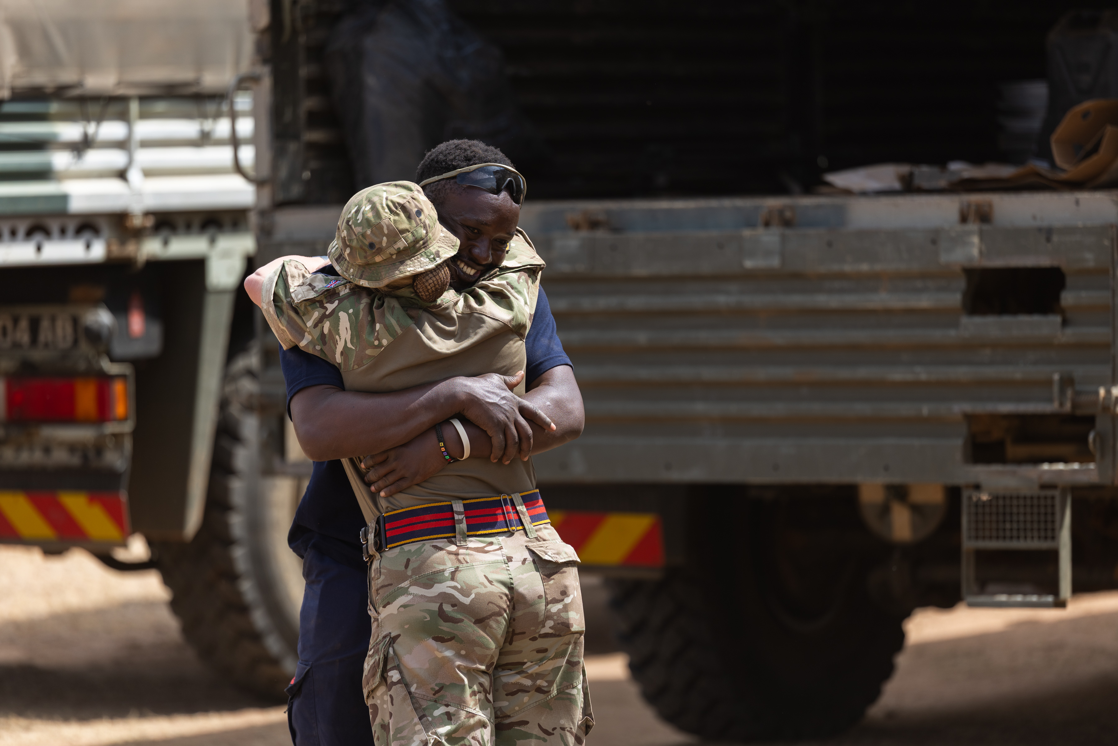 A soldier in camouflage uniform embraces a man wearing dark clothing near military trucks in a warm reunion.