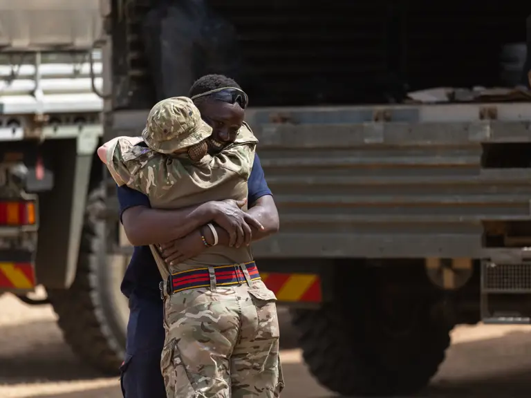 A soldier in camouflage uniform embraces a man wearing dark clothing near military trucks in a warm reunion.