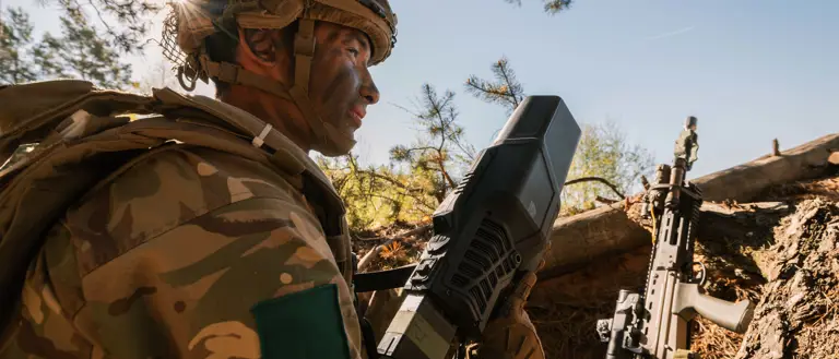 Soldier in camo gear, holding a large device, crouches in a forest setting near a rifle. Sunlight filters through branches, conveying tension and focus.