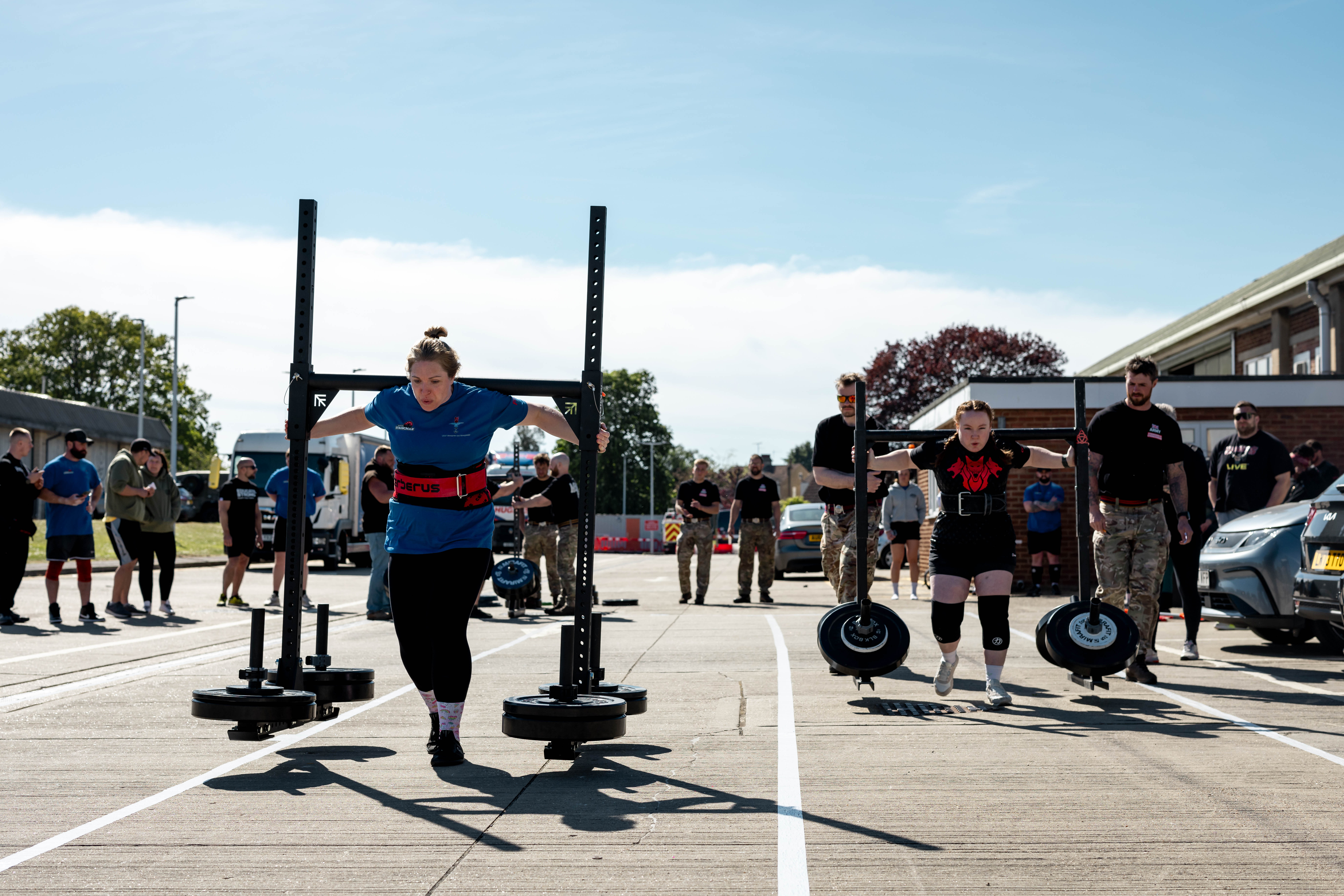 Two women compete in an outdoor strongman event, lifting heavy yokes. Spectators watch under a clear sky, creating a focused and intense atmosphere.