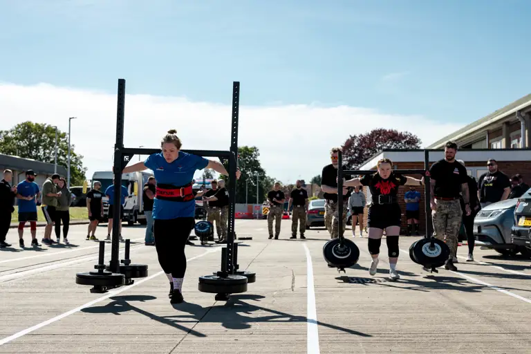 Two women compete in an outdoor strongman event, lifting heavy yokes. Spectators watch under a clear sky, creating a focused and intense atmosphere.