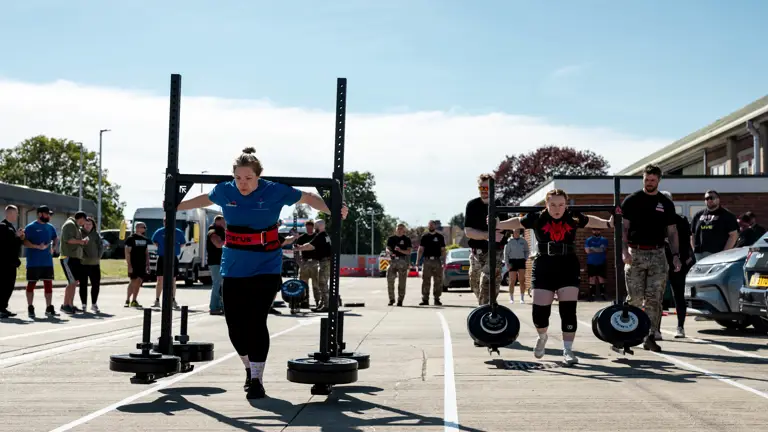 Two women compete in an outdoor strongman event, lifting heavy yokes. Spectators watch under a clear sky, creating a focused and intense atmosphere.