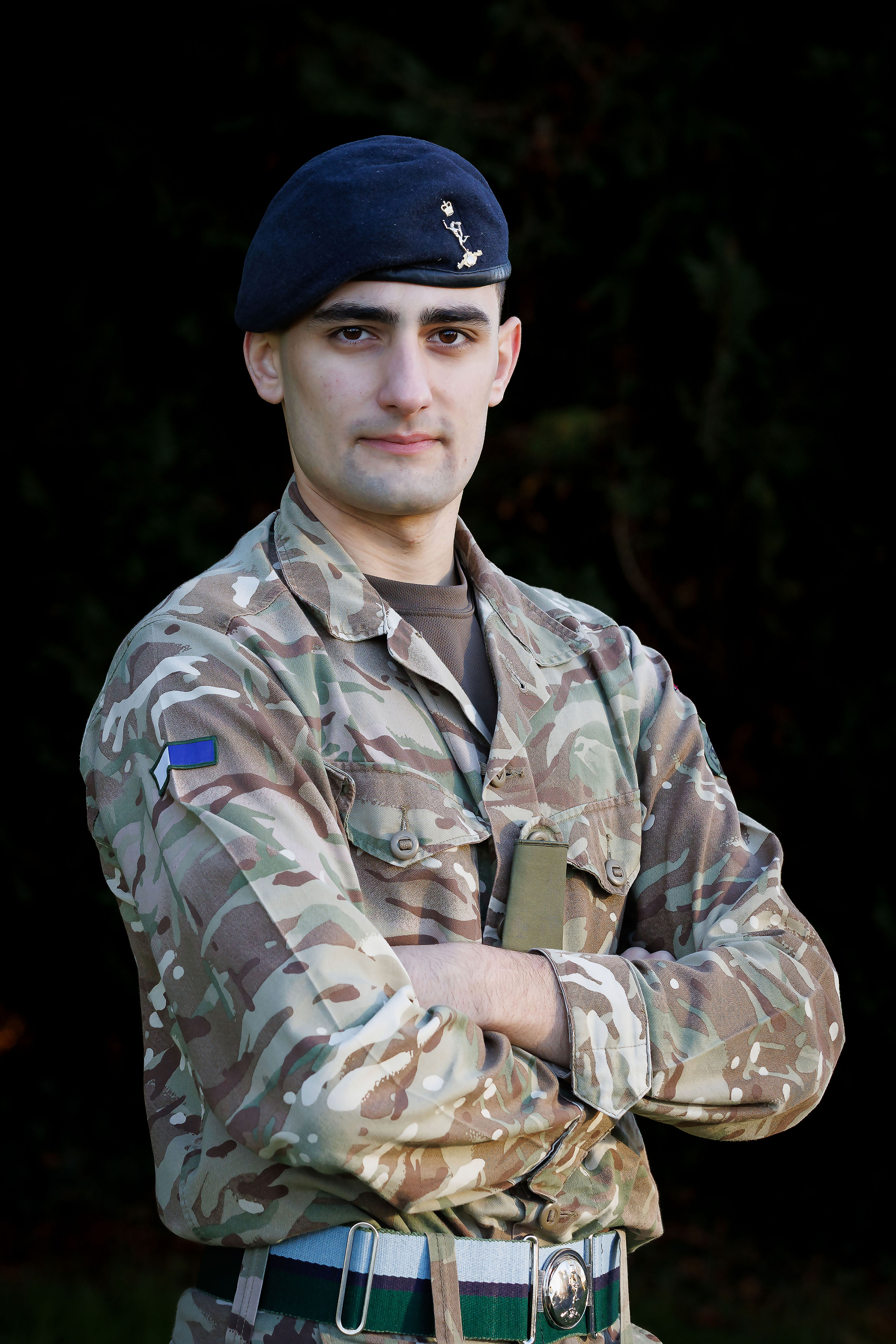 Soldier in camouflage uniform and beret stands confidently with arms crossed against a dark backdrop, exuding strength and determination.
