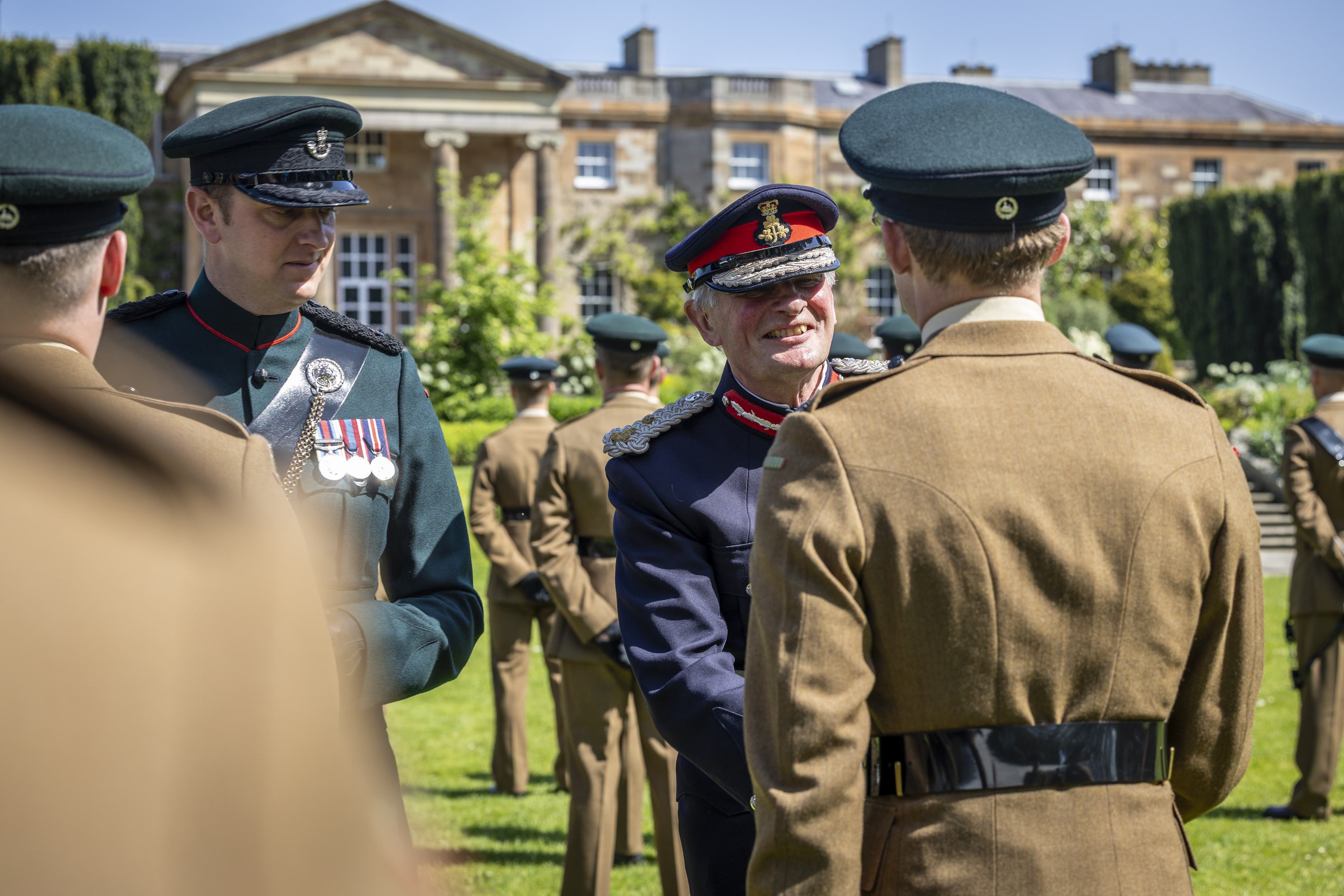 A group of uniformed individuals is outdoors, with one person in a decorated uniform smiling and shaking hands with another. In the background, there is a large, stately building. The scene suggests a formal military or ceremonial event.