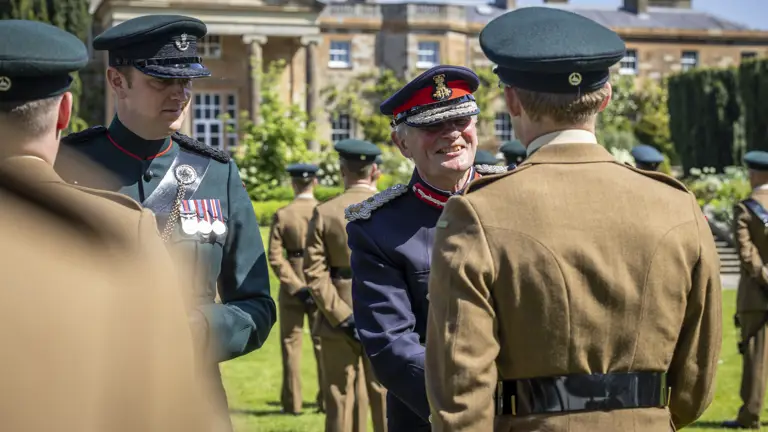 A group of uniformed individuals is outdoors, with one person in a decorated uniform smiling and shaking hands with another. In the background, there is a large, stately building. The scene suggests a formal military or ceremonial event.
