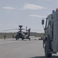 Image shows an AH64E Apache Helicopter stationary on the ground being refuelled by army air corps soldiers. A large Army vehicle is positioned on the right in the foreground.